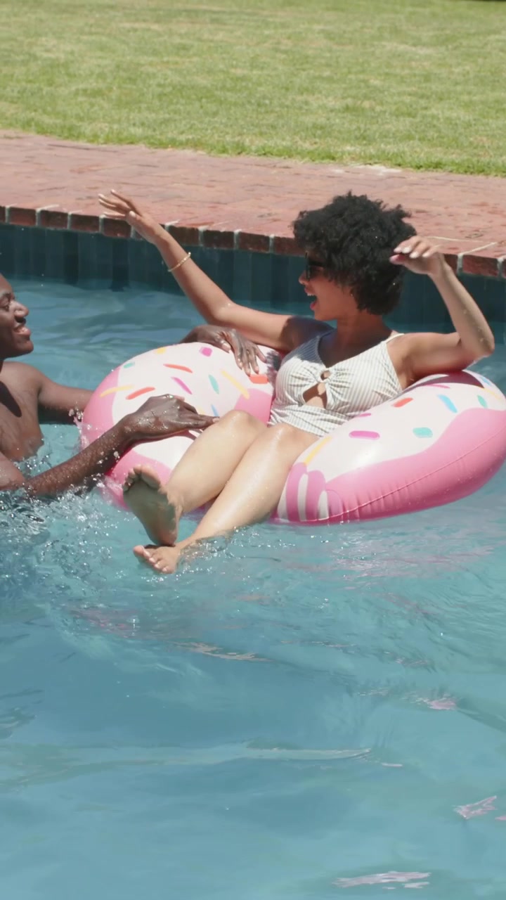 Vertical video: Floating woman in white swimsuit pool, man entering left steadying pink donut float
