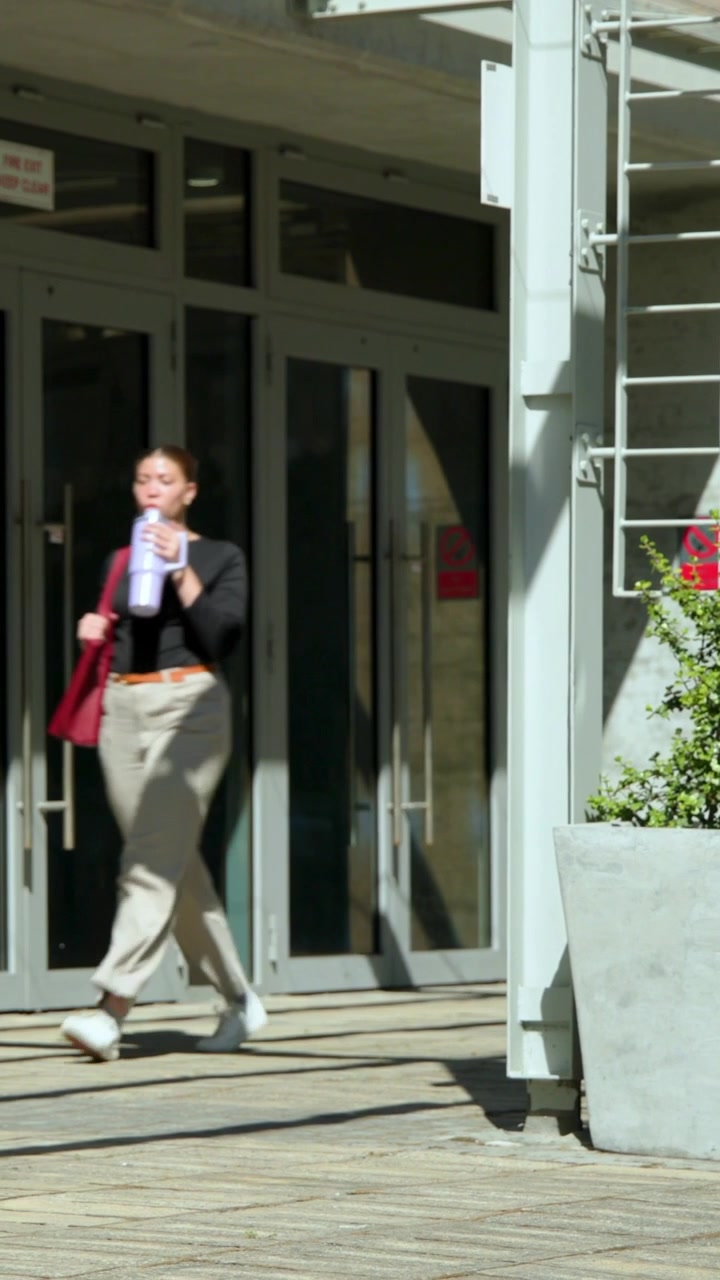 Video verticale: Donna che esce con una borsa rossa, passando una fioriera all'ingresso dell'edificio, lasciando l'edificio.