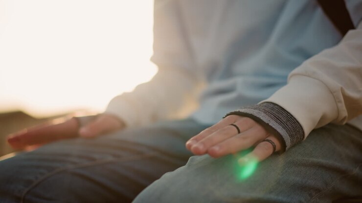 primo piano di una mano con anello su jeans, controluce dorata e un leggero flare dell'obiettivo che illuminano le dita in posa; la scena del parco urbano mostra un musicista paziente che tocca terra e prepara un set acustico con uno stile casual