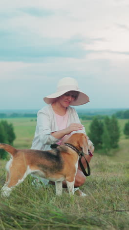 mujer sonriente con sombrero de sol agachada al lado de su perro feliz, tocando suavemente su cabeza mientras sostiene la correa, el fondo presenta campos verdes y árboles exuberantes