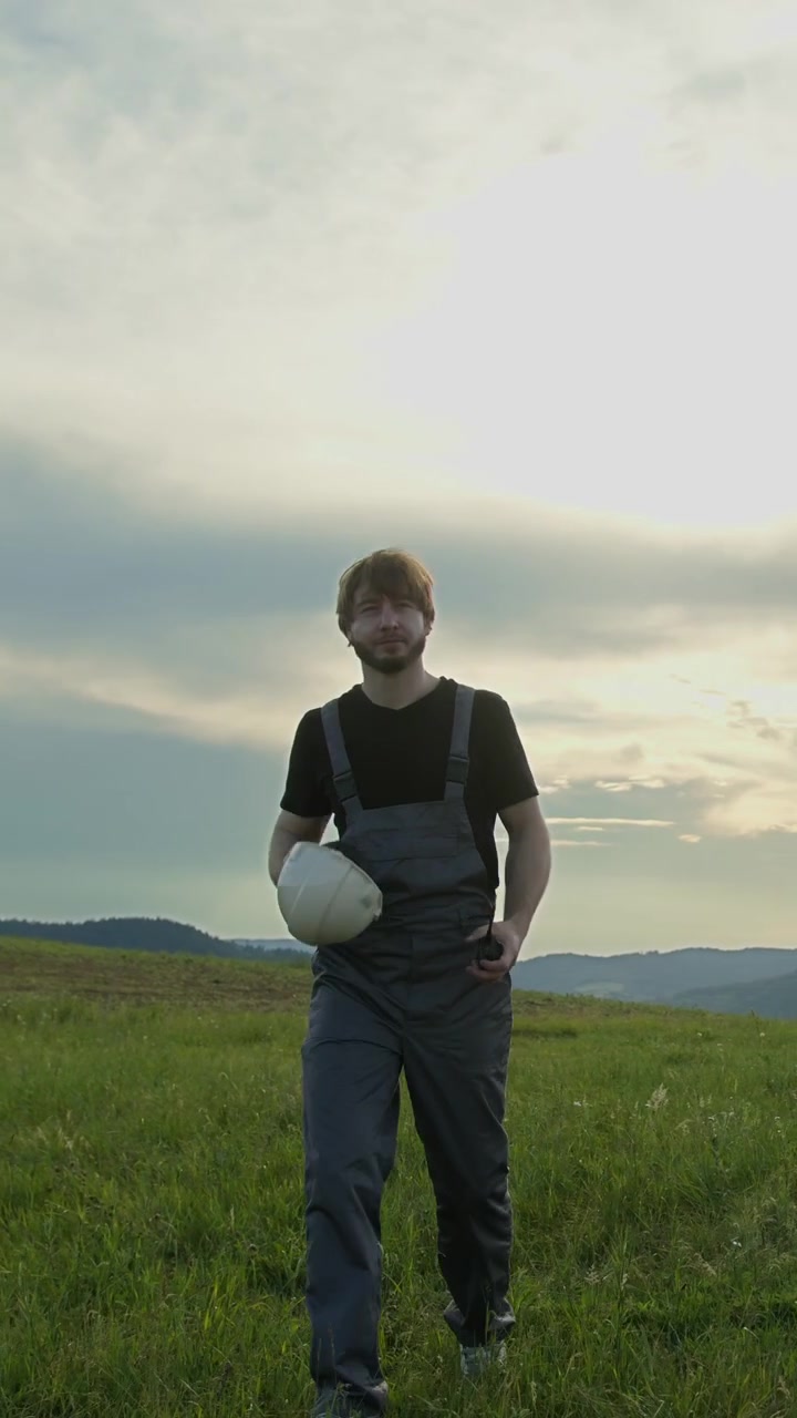 Uomo in tuta da lavoro e cappello cammina in un campo erboso vicino a turbine eoliche al tramonto.