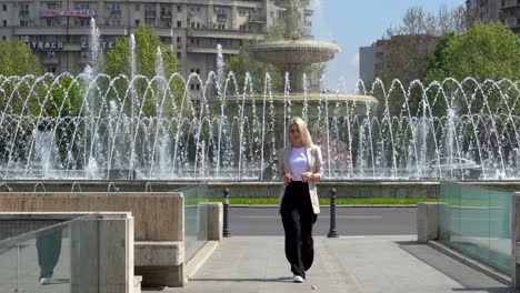 blonde young woman walking towards the camera with a water fountain in the background and passing cars, unirii square, bucharest, romania