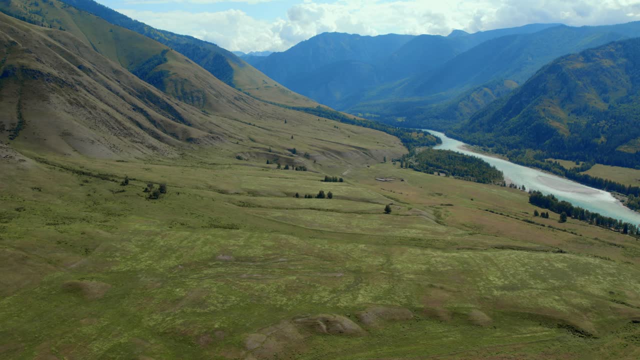 Riprese aeree con drone che sorvolano la Valle di Ak-Kem sui Monti Altai, Russia, mostrando un fiume serpeggiante, pendii verdi della steppa e cime aspre sotto un cielo nuvoloso.