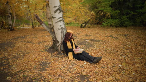 young lady in yellow beret sits outdoors, leaning against tree, flipping through book surrounded by dry autumn leaves, enjoying peaceful solitude in nature, lost in thought, immersed in reading