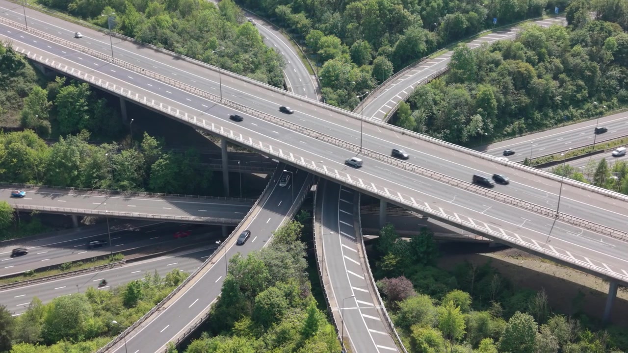 Cars, trucks, and a bus move through a sprawling, multi-level highway interchange. Elevated roads and ramps connect a complex network surrounded by green trees under clear daytime light.
