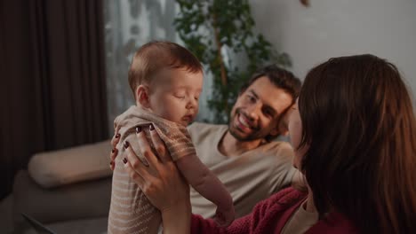 Happy-young-brunette-mother-in-a-red-sweater-together-with-her-brunette-husband-with-her-little-daughter-as-a-baby-and-looking-at-her-in-a-modern-apartment