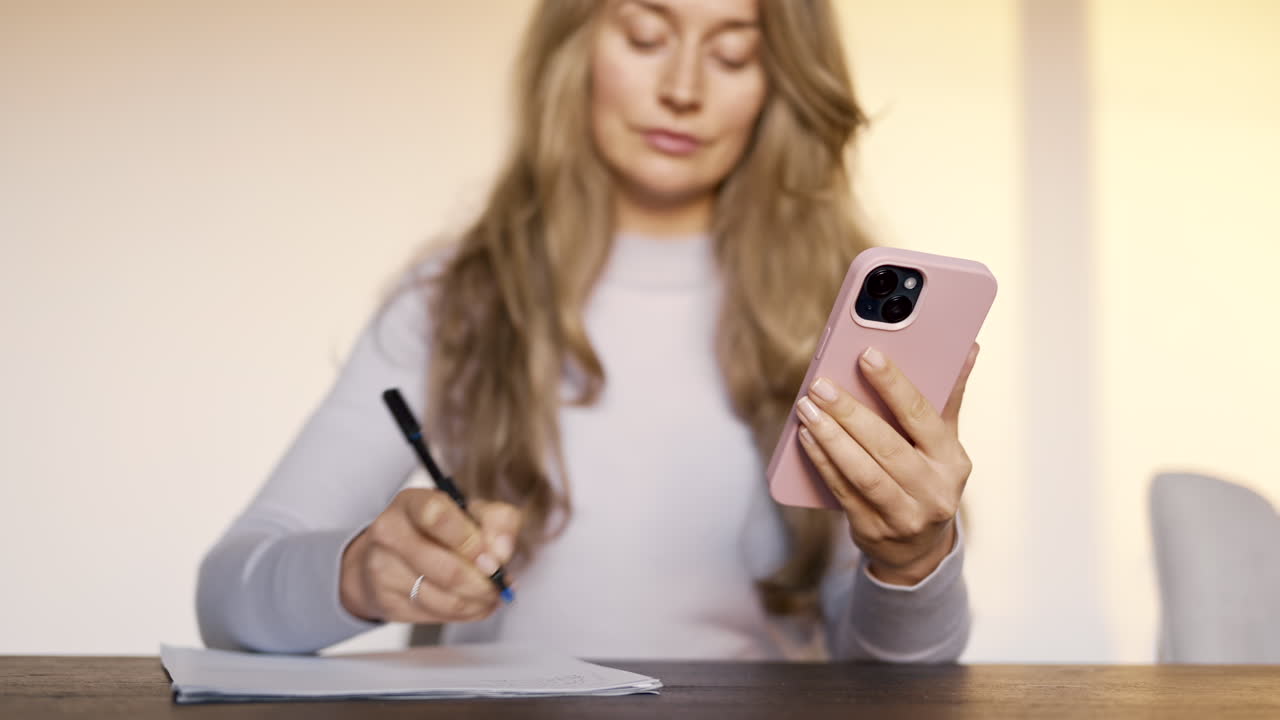 Woman writing on a piece of paper on a table while holding a phone
