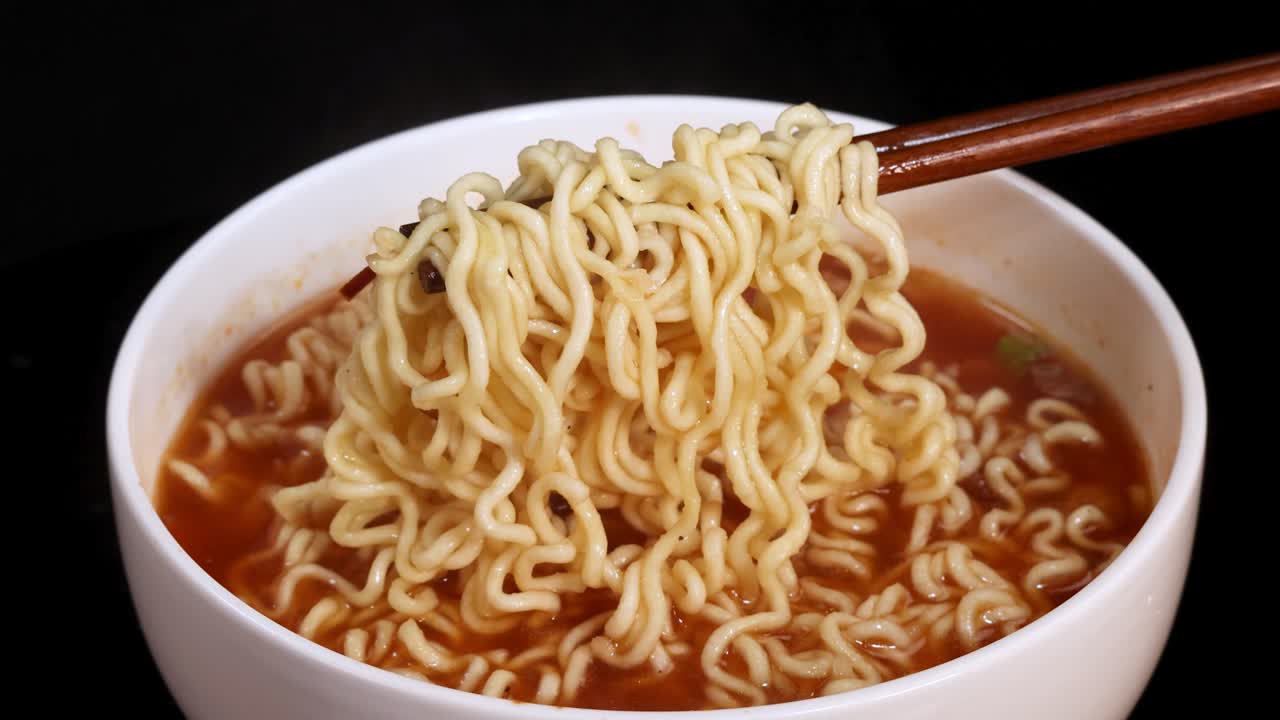 Chopsticks lift a portion of cooked instant noodles from a steaming bowl of broth under bright, even lighting, with a static, close-up camera angle