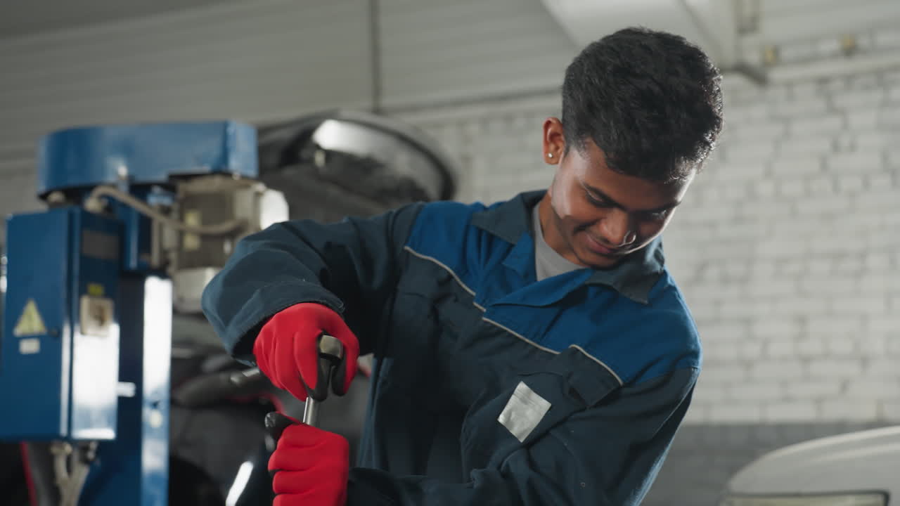 ingeniero automotriz con uniforme de trabajo azul y guantes rojos sonriendo mientras repara el motor del automóvil en el taller, el fondo presenta herramientas industriales, capó abierto del automóvil y equipos mecánicos