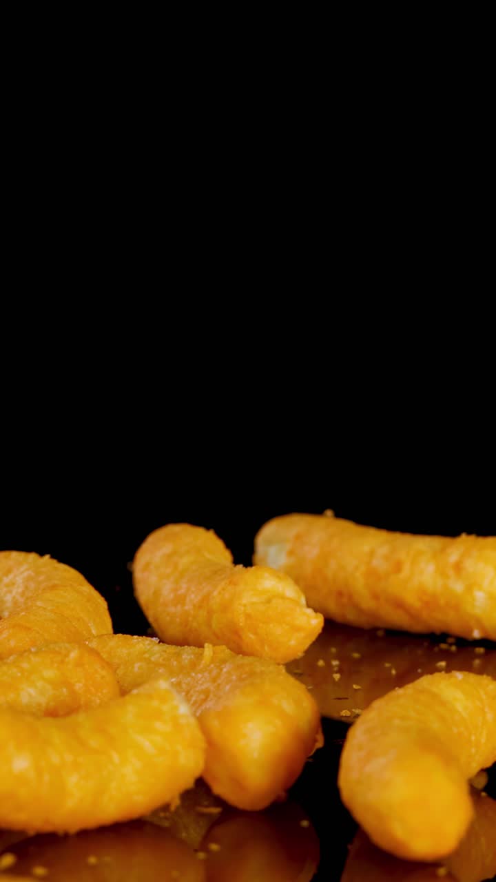 Puffs de queijo crocantes caem e se acomodam sobre um fundo preto brilhante, iluminação dramática, em close-up.