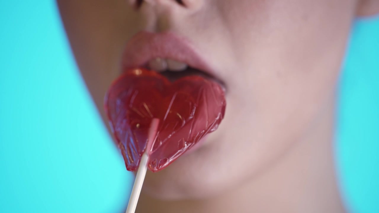 Macro ripresa 4K in slow motion di una donna che lecca un lecca-lecca a forma di cuore, sfondo blu vibrante, atmosfera romantica e giocosa di San Valentino, dettaglio ravvicinato, estetica cinematografica con effetto lucido.