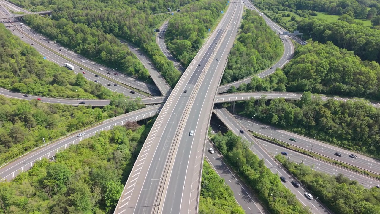 An aerial perspective shows vehicles navigating a complex motorway interchange, known as a spaghetti junction, surrounded by dense green forest during daytime.