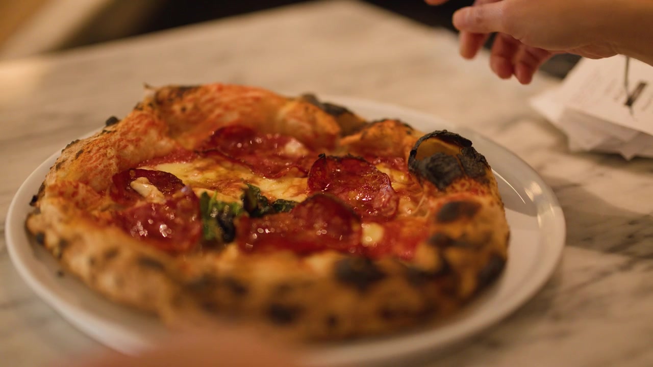 Primo piano di una mano che sistema un piatto con pizza al salame piccante con impasto a lievitazione naturale su un tavolo di marmo