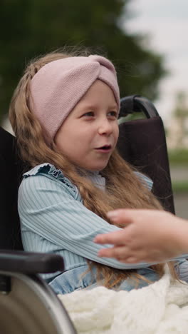 curly-haired girl enjoys playing fun hand games with mother in city park. woman entertains little daughter with leg injury in wheelchair closeup