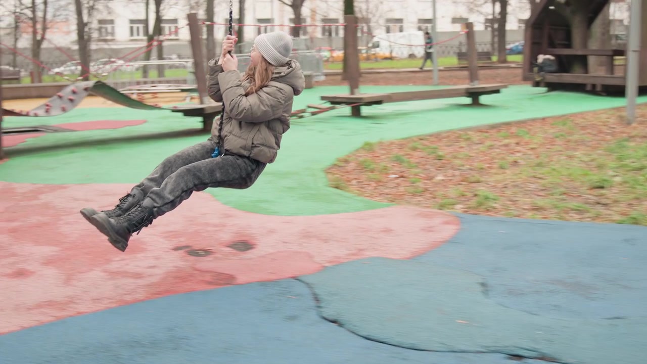 Woman on playground zipline under wooden arch, winter coat and beanie, hands gripping rope, cautious excited expression, urban park with bare trees, soft overcast light, solo adult exploring childhood