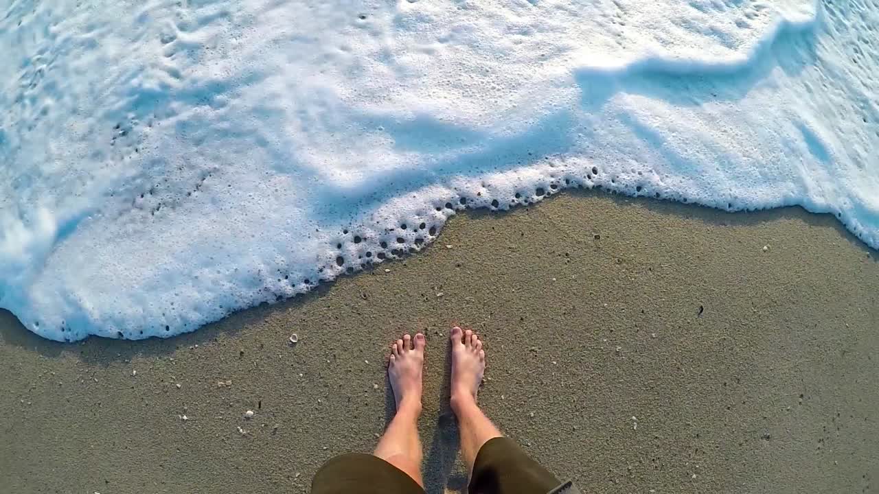 Male standing on a sandy beach and letting the waves roll up to his feet.