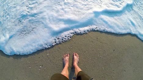 male standing on a sandy beach and letting the waves roll up to his feet.
