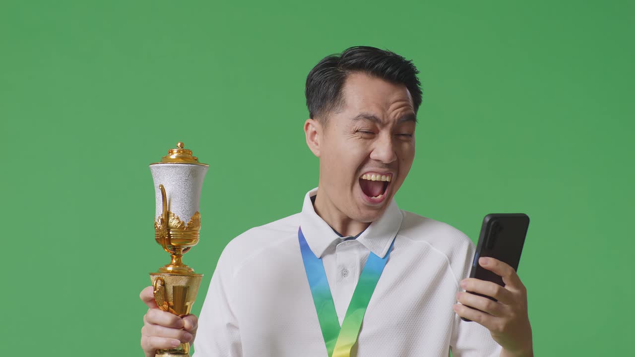 금메달과 트로피를 들고 스마트폰을 바라보는 아시아 남자의 클로즈업, 그 다음에는 스튜디오의 녹색 스크린 배경에서 축하하는 골을 비명을 지르고 있습니다.