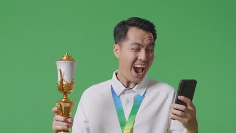 close up of asian man with a gold medal and trophy looking at smartphone then screaming goal celebrating on green screen background in the studio