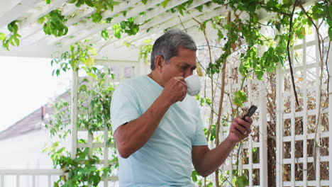 Hombre-Mayor-De-Raza-Mixta-Bebiendo-Café-Usando-Un-Teléfono-Inteligente-En-El-Jardín