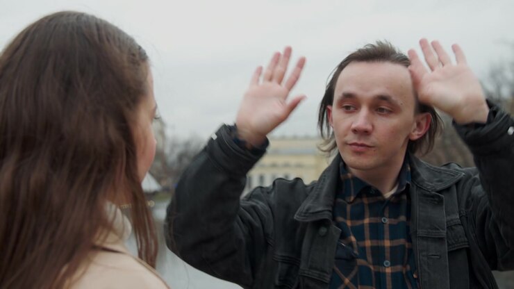 Primo piano di due persone all'aperto vicino a uno specchio d'acqua e a edifici. Una donna con i capelli lunghi tocca la giacca di un uomo mentre conversano, entrambi facendo gesti espressivi con le mani.