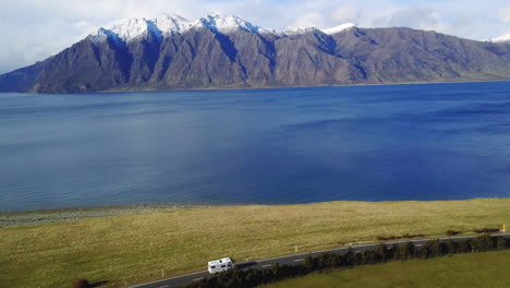 aerial video of a van driving on a coastal road with green paddocks, then panning to the blue ocean, and snowy mountains