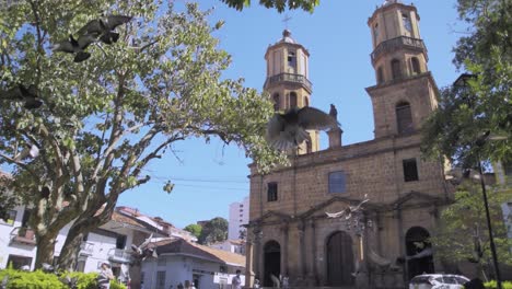 Der-Zentrale-Park-Von-San-Gil-Santander-Kolumbien,-Während-Tauben-In-Der-Mitte-Des-Platzes-Fliegen---Mit-Der-Stadt-Und-Der-Kathedrale-Dahinter---Schwebende-Taubenschwärme---Herumfliegende-Taubenschwärme