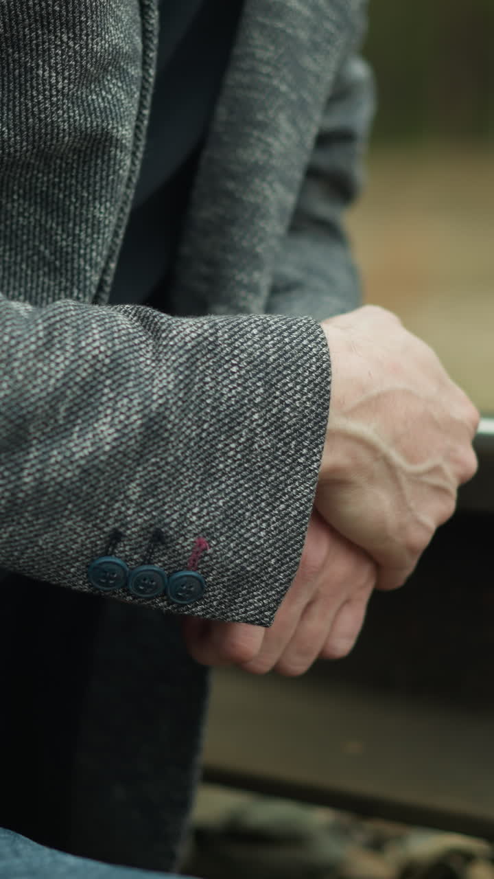 A close-up view of a man wearing a grey blazer and blue jeans, sitting and clasping his hands together on a railway track