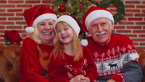 Senior-grandparents-with-granddaughter-in-Santa-hats-laughing-out-loud-at-home-near-Christmas-tree