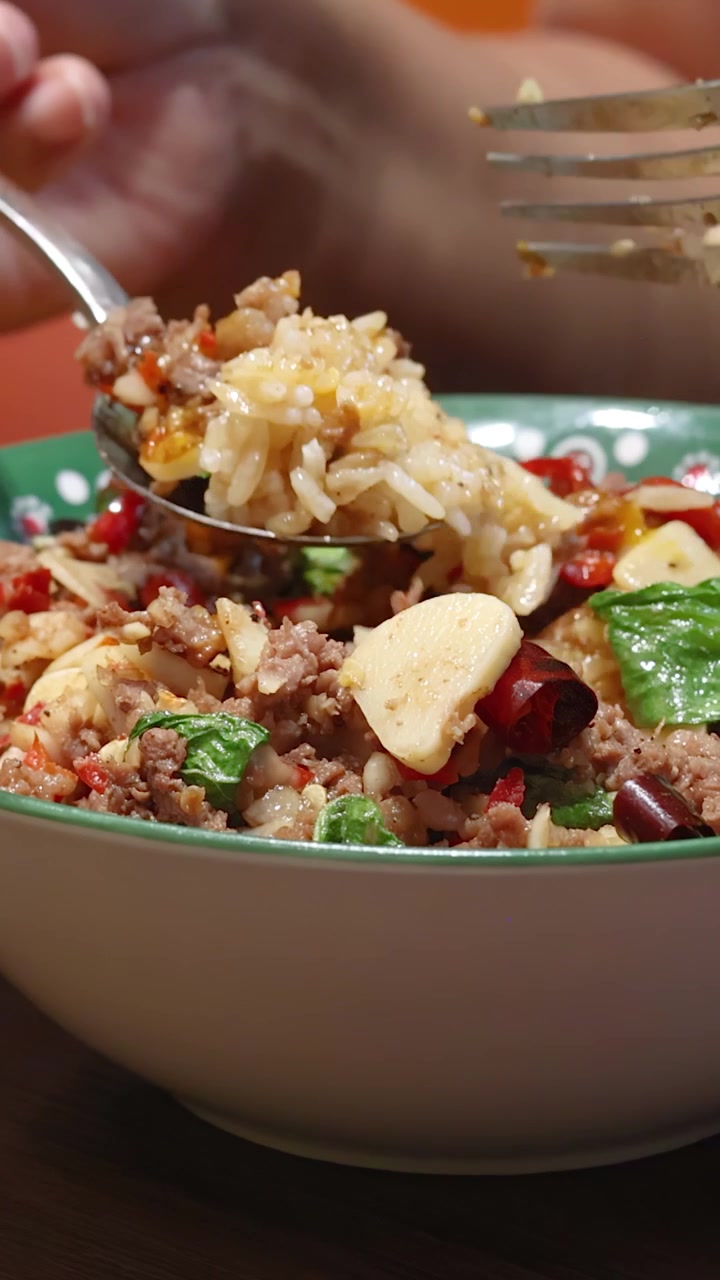 Una persona usa un cucchiaio e una forchetta per raccogliere manzo piccante, aglio, peperoncino e riso da una ciotola colorata sotto una calda illuminazione interna, vista ravvicinata.