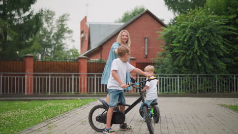 a mother is seen interacting with her children on a paved path in front of a house, she extends her hand, telling her older child to wait as she gives him a high five