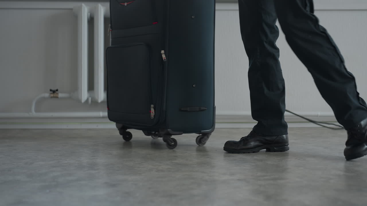 Tourist walks away from window with large wheeled travel bag on indoor concrete floor, black pants and shoes visible, radiator and residential building exterior faintly seen through window area