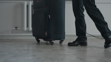 tourist walks away from window with large wheeled travel bag on indoor concrete floor, black pants and shoes visible, radiator and residential building exterior faintly seen through window area