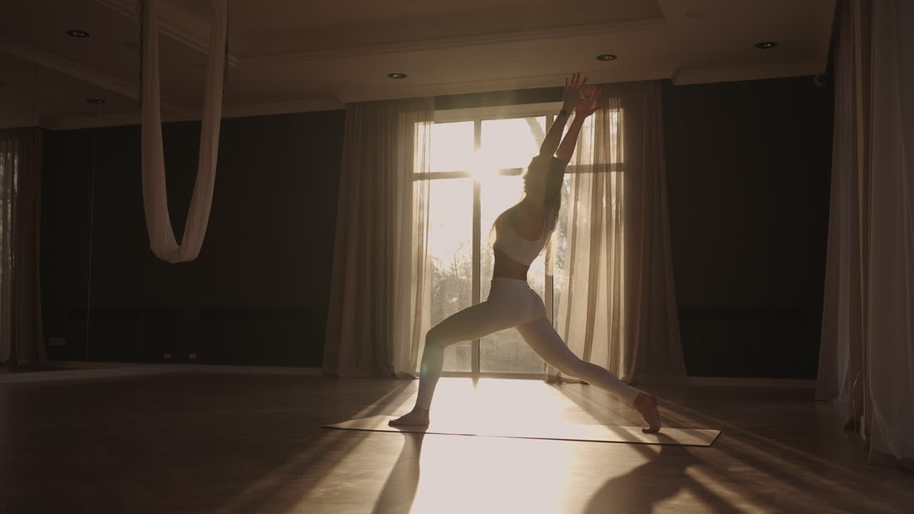 Slow motion: Young woman is doing yoga in a white room filled with light the girl performs yoga stands and elements near the large window.