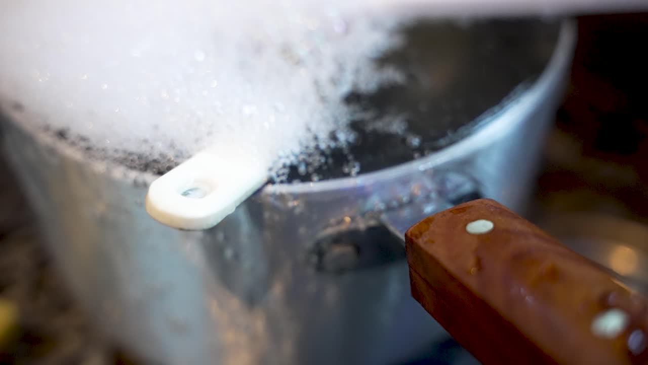 A silver pot sits in a kitchen sink overflowing with soapy water and bubbles with water pouring down the side