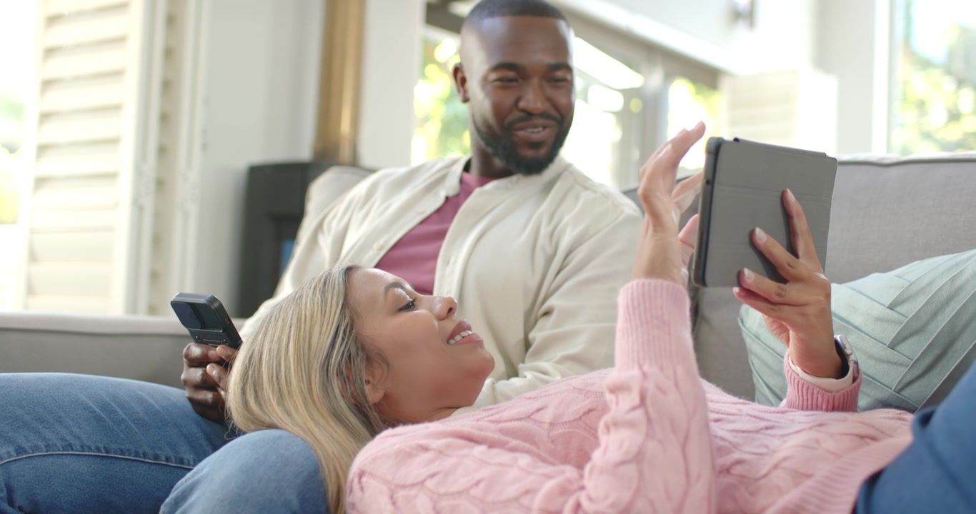 Coppia afroamericana che si rilassa sul divano, la donna tocca il tablet, l'uomo alza lo sguardo dal telefono sorridendo.