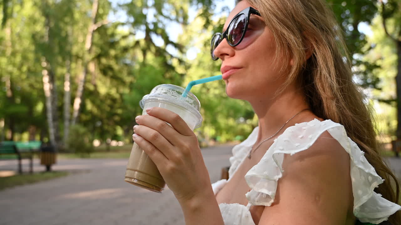 Brunette woman in white dress sipping an iced latte in a park