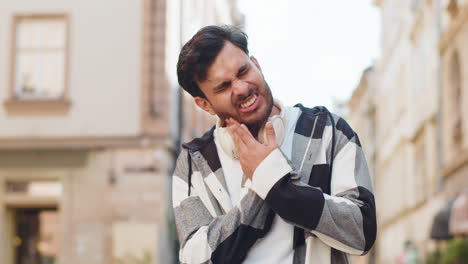 upset hispanic adult young man traveler feels a toothache while standing in urban city street