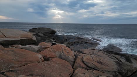 Vista-Aérea-De-Una-Mujer-Caminando-Y-Escalando-Rocas-En-La-Costa-De-Australia-Durante-La-Puesta-De-Sol-Con-Nubes-En-El-Cielo