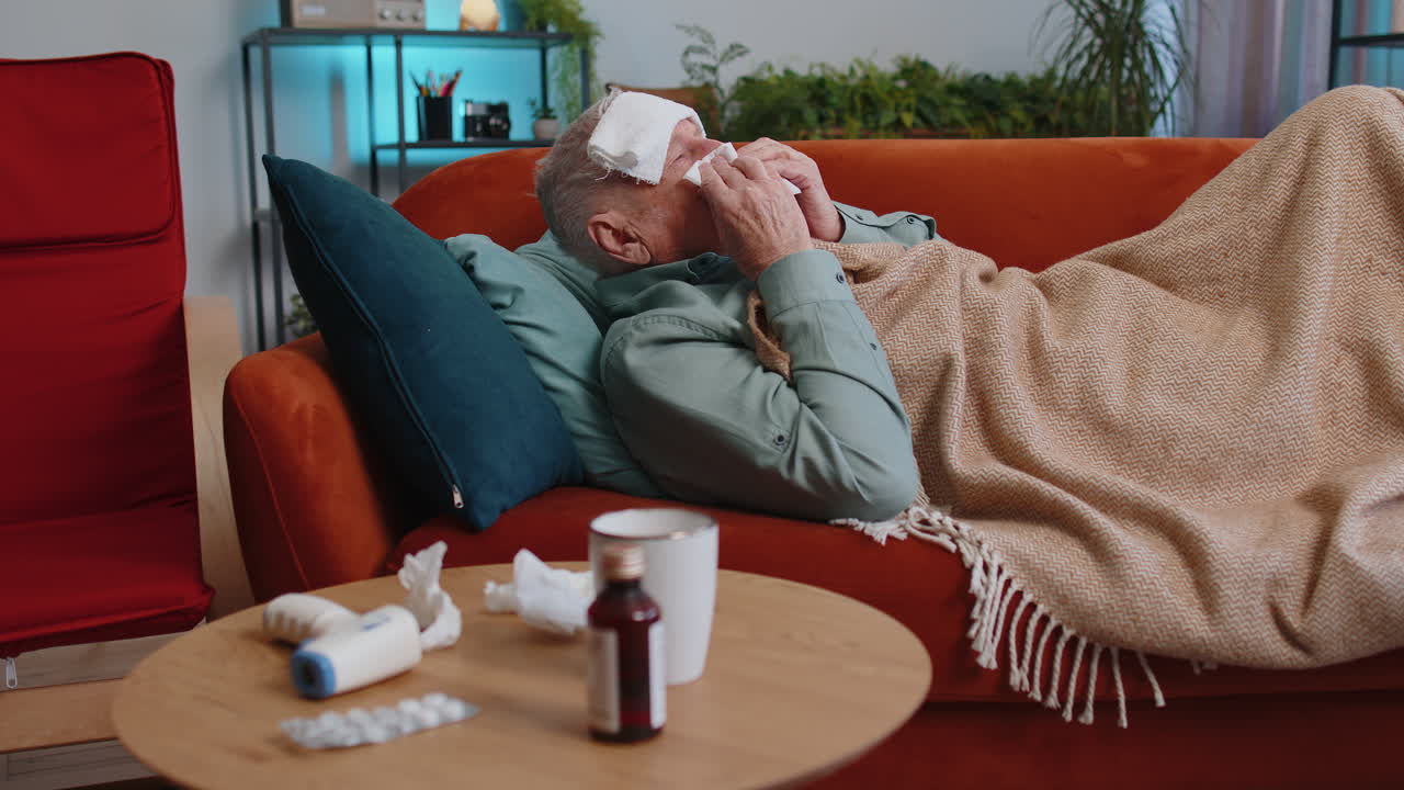 Sick ill senior man suffering from cold or allergy lying on home sofa sneezes wipes snot into napkin