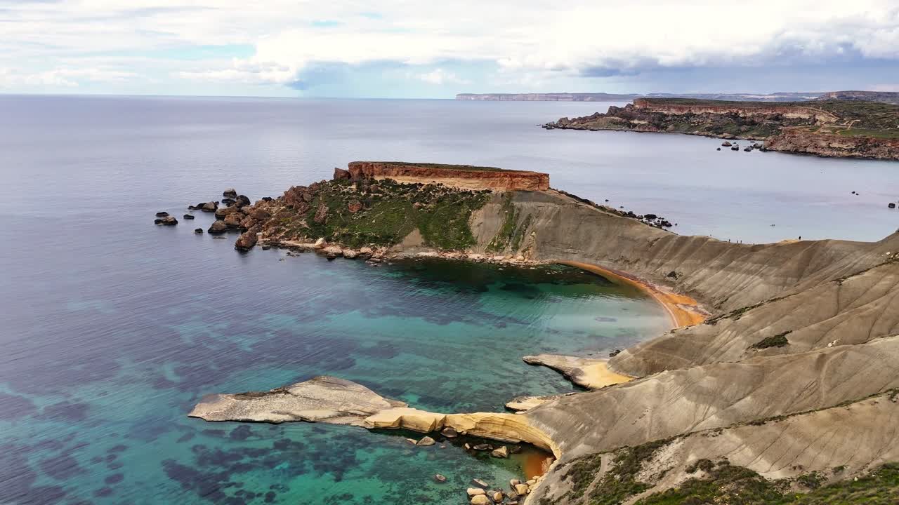 Baía de Qarraba em Mġarr, Malta, com encostas de argila azul sob um planalto de calcário, formando uma enseada curva sobre águas claras do Mediterrâneo dentro do sistema da Baía de Ġnejna, tomada de drone com aproximação e descida.