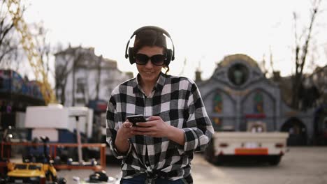 trendy, brunette in sunglasses walking on the street and listen to the music in headphones. dances. holding mobile in hands and searching for something. sunny day