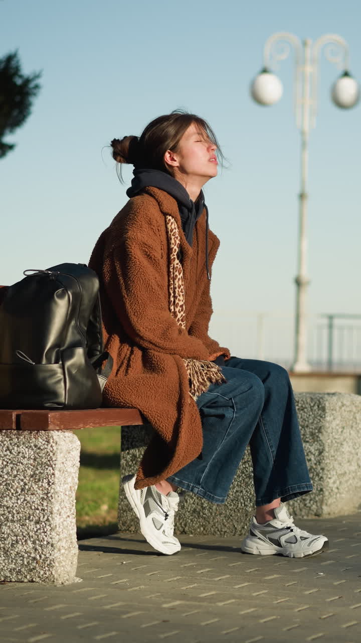 한 소녀가 도시 공원의 벤치에 혼자 앉아, 눈을 감고 생각에 빠져, 갈색 코트, 파란 청바지, 색 신발을 입고, 에 검은색 배을 고 있다.