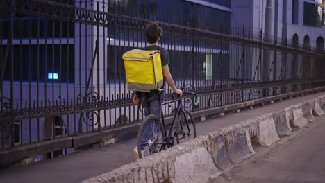 Delivery-man-walking-with-bike-and-yellow-bag-by-city-bridge,-rear-view