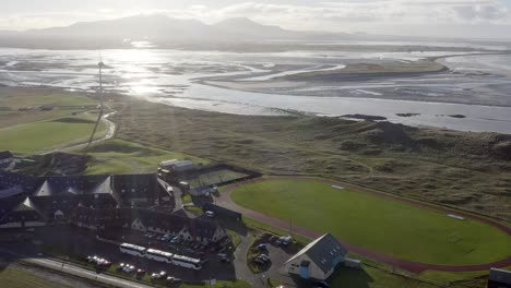 Reversing,-wide-angle-drone-shot-of-the-sea-and-coastline-around-Benbecula,-featuring-the-local-wind-turbine-and-football-players-playing-on-the-local-track-and-pitch