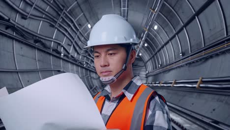 close up side view of asian male engineer with safety helmet looking at blueprint in his hands and looking around while standing in underground subway tunnel