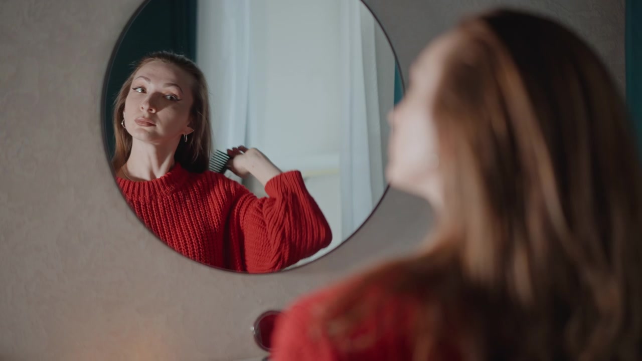 Giovane donna con un maglione rosso si china verso uno specchio rotondo, sistemandosi i capelli, toccandosi un orecchino e controllando il viso con un'espressione concentrata in una morbida luce interna durante la sua routine di cura personale.