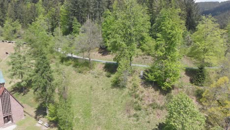 Aerial-view-panning-right-over-a-stone-church-in-a-forest-with-hiking-trails-on-the-mountains-as-people-walking-by