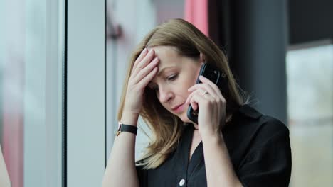 a stressed woman in black holds her phone and leans against a window in daylight