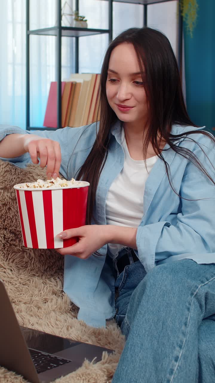 Woman happily eating popcorn watching movie on laptop at home sofa emotionally laughing enjoyment